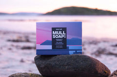 an island lavender soap box sitting on a rock at a white sandy beach. The box is a purple seascape design. The soap is described as soothing and calming. Handmade in Tobermory by the Isle of Mull soap co.