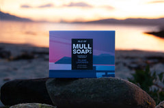 an island lavender soap box sitting on a rock at a white sandy beach at sunset The box is a purple seascape design. The soap is described as soothing and calming. Handmade in Tobermory by the Isle of Mull soap co.