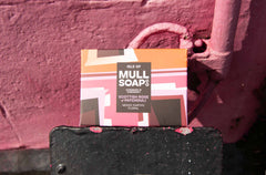 a Scottish rose and patchouli soap box displayed next to a sunny wall painted pink. The box is a pink colourful design. The soap is described as mossy, earthy and floral. Handmade in Tobermory by the Isle of Mull soap co.