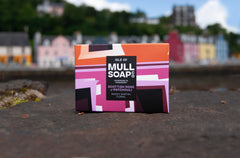 a Scottish rose and patchouli soap box displayed on Tobermory pier. The box is a pink colourful design shown infront of the colourful Main Street of houses and shops.. The soap is described as mossy, earthy and floral. Handmade in Tobermory by the Isle of Mull soap co.