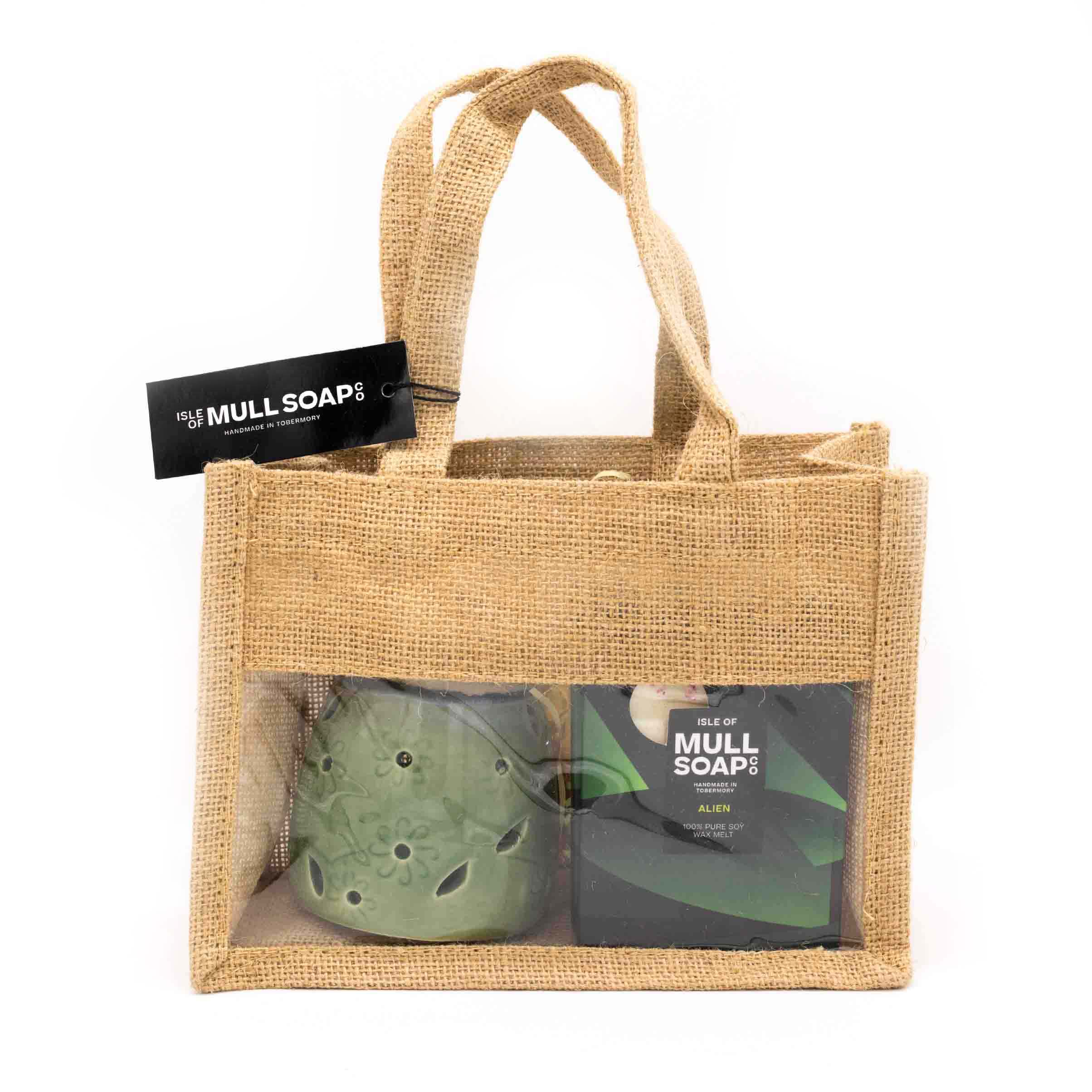 A natural coloured jute bag gift set with front window. Contains a green leaf and flower cut out ceramic burner and 2 Alien perfume scented soy wax melt snap bars made by the Isle of Mull soap co in Tobermory. contains packing shred and handles tied with a black tag.