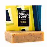 a vegan honey and oatmeal soap box sitting on top of the brown soap bar. The box is a blue and yellow design. The soap is described as exfoliating sweet and warm. Handmade in Tobermory by the Isle of Mull soap co.