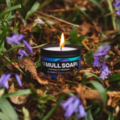 a lit bluebell soy wax candle with burning flame. nestled on a forest floor amongst dry leaves and bluebell flowers. handmade in Tobermory by the Isle of Mull Soap Co